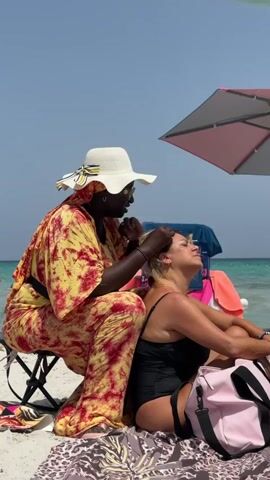 A person wearing a yellow and red tie-dye outfit and a white hat is braiding the hair of another person sitting on a beach. The person receiving the braid is wearing a black swimsuit and is leaning back with their eyes closed. A pink bag and an umbrella are visible in the background, along with the ocean and a clear sky.