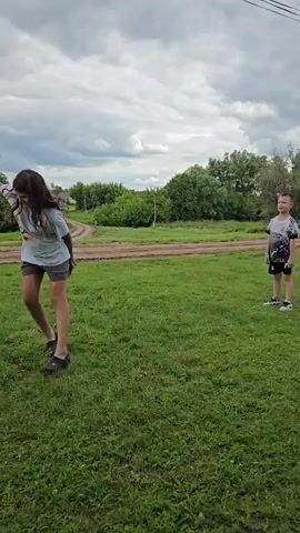 A young girl with curly brown hair, wearing a white t-shirt and patterned shorts, plays with a yellow soccer ball in a grassy field under a cloudy sky. She kicks the ball and then runs to catch it. Another young girl, wearing a pink jumpsuit, stands in the field and kicks the ball towards the first girl. The background shows a rural scene with houses, fences, trees, and power lines.
