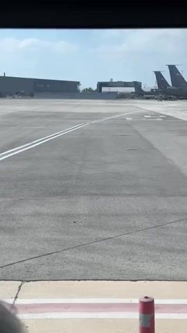 The video shows an aerial view of an airport tarmac. In the foreground, a grey tarmac is visible with white markings. In the background, several large, grey airplanes are parked. They appear to be military refueling aircraft due to their large size and the presence of a refueling boom extending from the wings of some of them. The planes are lined up neatly on the tarmac. Behind the airplanes, there are buildings and structures that suggest a military airbase or a large commercial airport. The sky is partly cloudy, indicating fair weather. The camera is moving, suggesting it is from a vehicle on the ground, possibly an airport bus or a follow-me car.