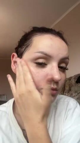 A young woman with dark hair pulled back, wearing a white t-shirt, is applying a clear gel or cream to her face. She is touching her cheek and forehead with her right hand. The lighting appears to be natural, coming from the side. The background is blurry but suggests an indoor setting with a wall and possibly furniture.
