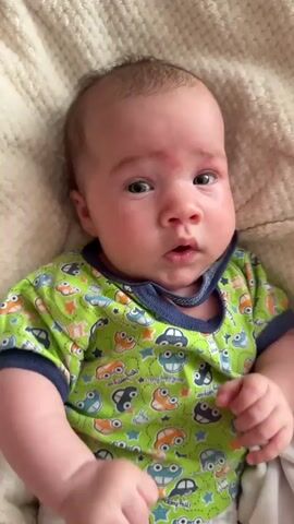 A close-up, high-angle shot shows a newborn baby lying on a cream-colored textured blanket. The baby is wearing a green short-sleeved onesie with a print of cartoon cars and stars. The baby's eyes are mostly closed at the beginning of the clip, but open and appear to look at the camera. A finger is visible entering the frame from the left side of the image and appears to be interacting with the baby. The baby's mouth is slightly open, and its face is red with what appear to be small red blemishes on its forehead. The lighting is soft and natural.
