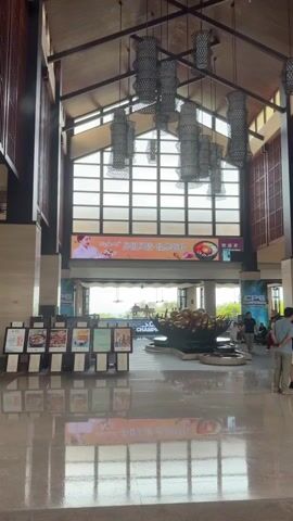 The video shows the interior of a large, open-plan lobby with a high, vaulted ceiling. Several large, decorative lanterns hang from the ceiling. A large LED screen is visible at the far end of the lobby, displaying text and images. In the foreground, there are several display stands with various posters or menus. The floor is polished and reflective. Towards the back of the lobby, there is a fountain with golden sculptures. A few people are visible in the lobby, some standing and some walking. The scene appears to be a hotel or a convention center. Outside the lobby, through large windows, there are buildings and palm trees visible. There are also cars parked outside. The lighting is bright, suggesting it is daytime.