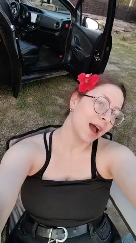 A young woman with blonde hair pulled back, wearing round glasses and a red flower in her hair, is sitting in a camping chair next to an open car door. She is wearing a black tank top and jeans. The scene appears to be outdoors, with grass and trees in the background, and a cloudy sky. The woman is looking at the camera and appears to be speaking. A second shot shows a different woman with blonde hair in sunglasses sitting on a blanket in a grassy field, with trees and power lines in the background.