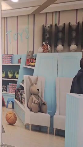 The video shows a toy store with shelves stocked with toys and stuffed animals. A large stuffed rabbit sits in a chair. Two young people are visible in the background, looking towards the toys. The store has a whimsical, colorful design.
