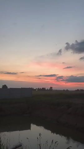 A video captures a serene natural landscape at dusk. The sky transitions from a light blue at the top to soft pinks and oranges near the horizon, painted with wispy clouds. A body of water, possibly a pond or a canal, occupies the foreground, its surface reflecting the muted colors of the sky. Along the bank of the water, there are mounds of soil and sparse vegetation. In the middle ground, a field of green grass stretches out, dotted with a few trees. On the left side, a portion of a gray, block-like structure is visible, possibly a wall or a building under construction. The overall impression is one of peaceful solitude in a rural setting as day turns into night.
