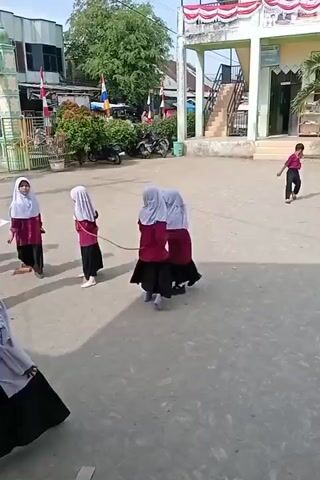 A group of at least five young girls, dressed in maroon shirts, black skirts, and white hijabs, are in an outdoor courtyard. They appear to be playing a game that involves a rope. One girl is swinging a rope, while others are positioned around her, seemingly preparing to jump or move. The ground is paved with gray concrete. In the background, there is a building with a green facade and a red and white awning, as well as trees, fences, and parked motorcycles. The sky is clear and bright.