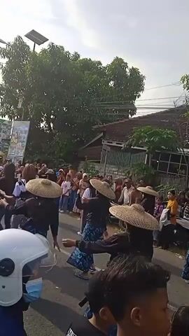 The video shows a crowded outdoor event, possibly a street parade or festival. Many people are present, including individuals wearing hijabs, some wearing traditional straw hats, and others dressed in casual clothing. Some people in the crowd are dancing, and there appears to be a stage or platform set up in the background with flashing lights and smoke effects. There is a building and tall pole visible near the crowd and it appears to be daytime with bright sunshine. The person filming is positioned in the crowd, occasionally looking up at the event.