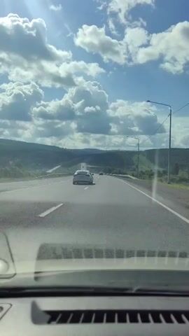 A POV shot from inside a vehicle driving on a highway. The road is asphalt with white lane markings. In the distance, a few cars are visible, including a white car in the right lane. The landscape on either side of the highway consists of rolling green hills and trees. The sky is blue with fluffy white clouds, and the sun is shining brightly, casting lens flares. The interior of the vehicle, including the dashboard and windshield, is visible in the foreground.
