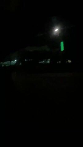 The video shows a dark outdoor scene at night. A large building or structure with a white roof is visible in the distance, lit by several interior lights. There are trees and some other structures in the foreground, also dimly lit. The perspective suggests the view is from a moving vehicle, as evidenced by the changing scenery and the blurred foreground. A street lamp with a green band is also visible. The overall ambiance is dark, with the lights from the building and street lamps serving as the primary sources of illumination. The video focuses on capturing the visual elements of the scene in low-light conditions.