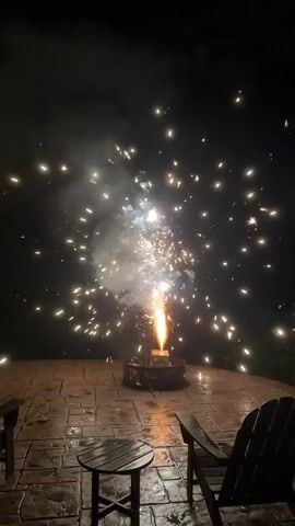 A nighttime video shows a small firework exploding on a patio. The patio is paved with stone and has two Adirondack chairs and a small round table. The firework is a fountain type, shooting sparks upwards and outwards in a fan shape. The firework is held in place by a small metal holder. The sparks are white and yellow, with some smoke visible. The background is dark, with trees visible on the right side of the frame. The patio appears to be wet, possibly from rain. The video is looped, with the same sequence of events repeating.