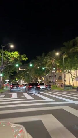 The video captures a nighttime scene on a city street, likely in a tropical location due to the presence of palm trees. The camera is positioned inside a vehicle, looking forward. Cars are traveling down the road, and a crosswalk with white stripes is prominently visible in the foreground. Traffic lights are illuminated green, indicating traffic is moving. Streetlights and lights from buildings illuminate the scene. Pedestrians are visible on the sidewalk to the right. The overall atmosphere is one of movement and urban activity at night.