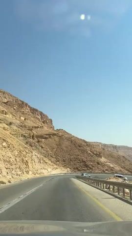 The video shows a view from a vehicle driving on a paved road. The road curves to the right and is bordered by a metal guardrail. To the left of the road, there is a steep, rocky, sandy mountain. The sky is clear and blue, with two bright sun flares visible. There are a few cars visible in the distance on the road.