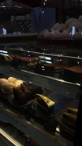 The video shows a display case in what appears to be a bakery or cafe. The display case is filled with various desserts, including rocky road, apple crumble tart, Italian cream buns, assorted brownies, and other pastries under glass domes. The lighting is somewhat dim, and the focus is on the food items displayed. There are labels indicating the names of the desserts and their prices. No people are visible in the frame.