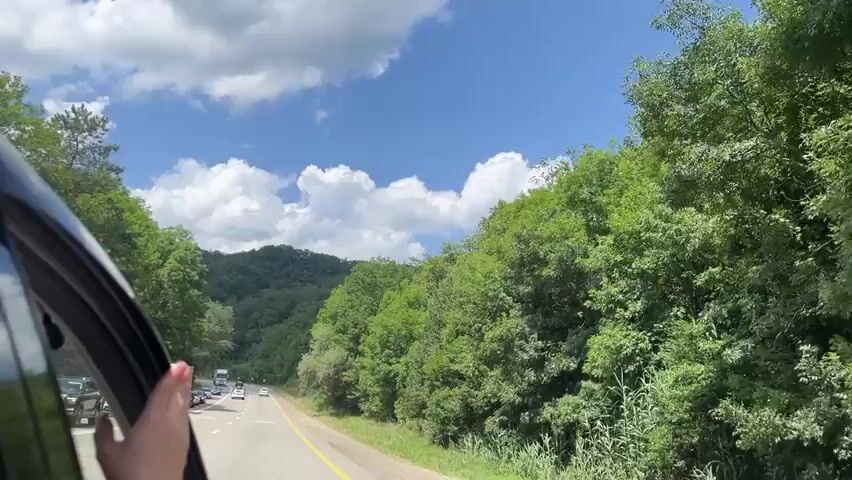 A first-person perspective video shows a view from inside a moving vehicle. The passenger's hand is visible in the lower left corner, resting on the open window frame. The road ahead is visible, with several cars and a truck in the distance. Lush green trees and rolling hills covered in dense forest line both sides of the road, extending into the background. The sky is blue with scattered white clouds. The overall scene depicts a scenic drive through a wooded, mountainous area on a sunny day.