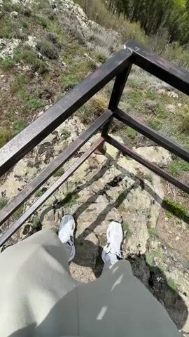 The video starts from a first-person perspective showing someone standing on a rocky ledge behind a black metal railing, wearing white sneakers and light grey pants. The camera pans up to reveal a panoramic view of lush green mountains under a sky dotted with fluffy white clouds. In the distance, a small town is visible nestled among the hills. As the camera pans further, it reveals a small, boxy structure covered with a tarp on the rocky peak. The scene is serene and showcases a natural, mountainous landscape.
