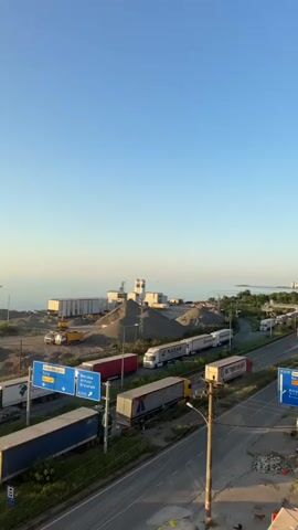 A vertical video shows a clear blue sky above a coastal industrial area. The foreground is dominated by a road with multiple lanes, bordered by a grassy median and a sidewalk. Several large semi-trailer trucks are visible on the road, some stationary and others appearing to be in slow-moving traffic. Blue directional signs are posted along the road, indicating routes. Beyond the road, there's a large open area with piles of gravel and construction materials, as well as shipping containers and industrial buildings. A yellow construction vehicle is partially visible. Further back, the sea is visible, with a calm surface and a cargo ship in the distance. The sun appears to be low in the sky, casting a warm light, suggesting either early morning or late afternoon. The overall impression is one of a busy port or logistics hub near the coast.