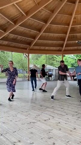 A group of people are dancing in a circular, open-air pavilion with a wooden roof and floor. The pavilion is supported by wooden beams. In the foreground, a woman in a dark blue dress with white polka dots is dancing, facing away from the camera. To her right, a man in a black t-shirt and white shorts is dancing. Further to the right, two couples are dancing. One couple consists of a man in a striped shirt and light-colored pants, and a woman in a blue t-shirt and black pants. The other couple is not fully visible. In the background, more people are visible, some standing and some dancing. The environment appears to be outdoors, with trees and other structures visible beyond the pavilion. The lighting suggests it is daytime.