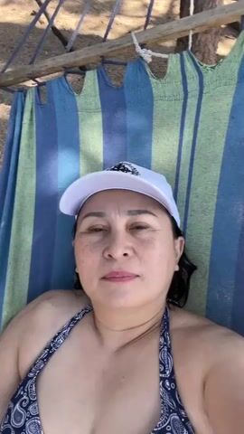 A medium close-up shot shows a woman lying in a hammock. She is wearing a white baseball cap and a blue and white patterned bikini top. The hammock has blue, green, and yellow stripes. The background appears to be outdoors, with trees and sand visible. The woman is looking at the camera and smiling.