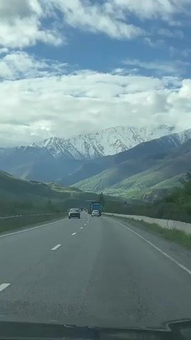 The video shows a car driving on a highway with a view of snow-capped mountains in the distance. The sky is blue with scattered clouds. There are other vehicles on the road, including a dark-colored car and a blue truck. The highway is paved and has lane markings. The surrounding landscape consists of green hills and trees. The video is taken from the perspective of someone inside the car.