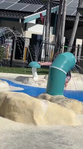 A child is playing in a splash pad. The child is wearing a long-sleeved blue shirt and patterned pants. The child is walking on the splash pad, which has blue water and sandy-colored rocks. There are mushroom-shaped water features in the background, one of which is green and is spraying water. Another mushroom-shaped feature is white and blue. The background shows a building with solar panels on the roof and palm trees. There is a black fence behind the splash pad.