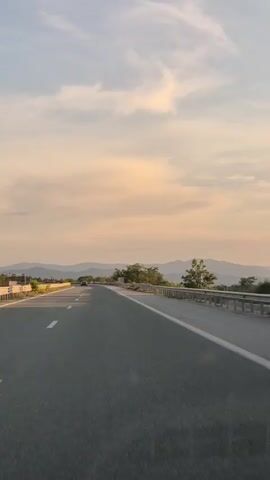 The video shows a point-of-view shot from a moving vehicle on a paved road. The road curves to the left, with a guardrail on the right. In the distance, there are mountains and a light-colored sky with scattered clouds. There are trees and vegetation on the side of the road and on the distant hills. The sun appears to be low in the sky, casting long shadows. There is at least one other vehicle visible in the distance on the same road. The overall scene is a landscape view of a road trip during either sunrise or sunset.