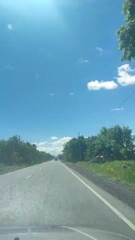 A dashcam footage captures an outward view from a vehicle traveling on a paved road under a bright blue sky with scattered white clouds. The sun is visible in the upper left corner, casting lens flare. Trees line both sides of the road. A white line marks the edge of the road on the right. In the distance, several vehicles are visible on the road. The lower portion of the windshield is visible at the bottom of the frame.