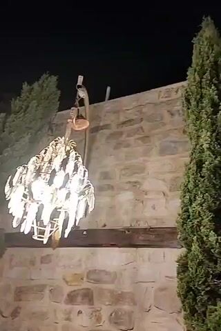 The video shows an outdoor scene at night, featuring a stone wall and tall evergreen trees. A chandelier hangs from a hook on the wall. The camera pans across the scene, capturing the texture of the stone wall and the silhouette of the trees against the dark sky. The chandelier is illuminated, casting a warm glow.