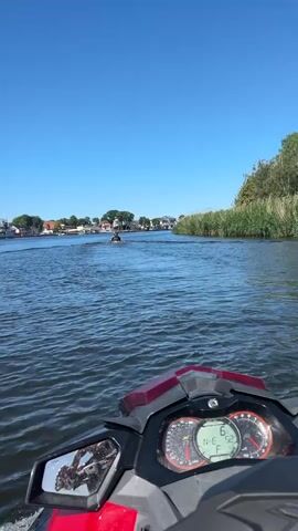 The video shows a first-person perspective from a jet ski on a clear, sunny day. The water is calm and blue, with a gentle ripple. In the distance, a town lines the shore, with buildings, trees, and a few boats docked. A military ship is visible in the background, suggesting the area may be a port or naval base. A second jet ski can be seen ahead, with two people on it. The camera pans slightly to the right, revealing a blonde woman with her back to the camera, wearing a life vest and looking out at the water. The time of day is clearly daytime, with a bright blue sky and strong sunlight, indicating warm weather. The jet ski's dashboard is partially visible, showing a speed of 6, possibly indicating 6 miles per hour or kilometers per hour, and a fuel gauge. The overall mood is one of leisure and enjoyment, with the activity being a recreational jet ski ride.