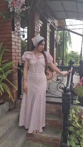 A young woman wearing a light pink floral dress and a white headscarf stands on a brick patio. She is leaning against a black metal railing. The woman is looking down and to her right. There are plants and flowers around her.
