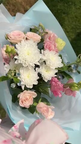 A close-up overhead shot shows a bouquet of flowers wrapped in light blue paper. The bouquet contains white chrysanthemums, light pink roses, pink ranunculus, and small blue flowers. The background is blurred, showing green grass. A portion of a light blue and pink object is visible at the bottom of the frame.