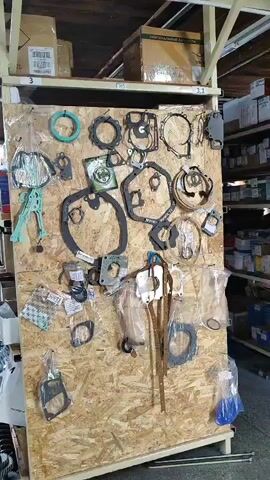 The video shows a large pegboard display filled with various gaskets. The pegboard is made of particle board and is set up in what appears to be a warehouse or parts storage area. Shelves filled with boxes are visible in the background. The gaskets are of different shapes and sizes, and some are still in plastic packaging. The lighting is somewhat dim, suggesting an indoor environment. There are no people visible in the video, and no actions or interactions are taking place. The overall scene is static and focused on the display of automotive or industrial gaskets.