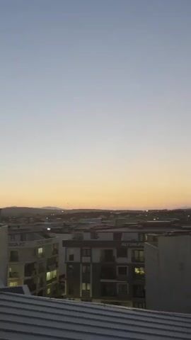 The video captures an aerial view of a city during twilight. The sky is clear and transitions from a pale blue at the top to a soft orange hue near the horizon. In the foreground, rooftops of buildings are visible, some with satellite dishes. The middle ground features apartment buildings with illuminated windows, indicating that the evening has settled. The background shows more of the city's urban landscape extending towards a distant, hazy mountain range under the fading light.