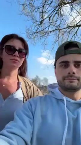 A woman in sunglasses and a beige jacket stands beside a man in a light blue hoodie and green baseball cap, who is holding the camera in selfie mode. The camera briefly shifts to reveal a third person, a young woman with glasses. They are outside, likely in a park or outdoor setting, with trees visible in the background under a partly cloudy sky. The man smiles as the camera captures the group.