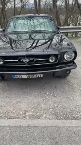The video shows a black 1965 Ford Mustang GT parked on a gravel road. The car is shiny and appears well-maintained, with a prominent Mustang emblem on the grill and a license plate reading "KR 1965 GT". The environment is outdoors, with bare trees visible in the background. The camera slowly pans to the left, offering a side view of the vehicle and its custom wheels. The scene is still and appears to be filmed on an overcast day.