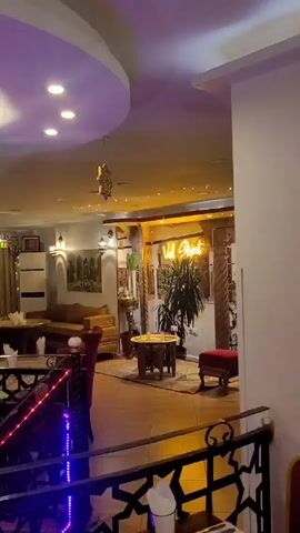 The video shows the interior of a restaurant with Moroccan-style decor. There are multiple seating areas with tables and chairs, as well as cushioned seating arrangements. The lighting includes overhead spotlights, colorful hanging lanterns, and decorative wall sconces. The restaurant has a two-level layout, with a railing separating the upper and lower sections, decorated with a geometric pattern and strung with colorful lights. The color scheme is warm, with earth tones and accents of red and gold. A neon sign with the word "guest" in Arabic script can be seen in the background.