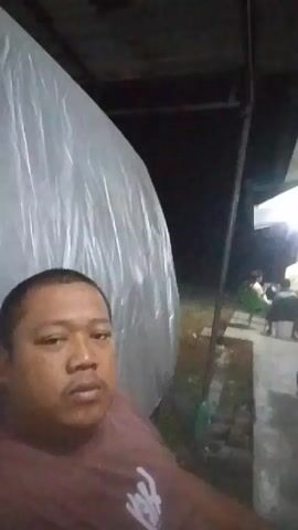 A man in a maroon t-shirt is filmed in a close-up shot. He is looking directly into the camera with a neutral expression. Behind him, out of focus, are several other people gathered around tables, suggesting an outdoor gathering. The environment appears to be dimly lit, possibly at night, with a large, translucent sheet or tarp dominating the left side of the frame. The overall impression is of a casual, informal setting.