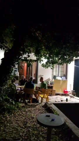 The video shows a group of four men sitting around a table outdoors at night. The men are casually dressed and appear to be engaged in conversation. The setting is dimly lit, with a house visible in the background. There are trees and foliage around the area, and a round table is in the foreground. The men are seated in chairs, with one man standing. The scene appears to be relaxed and social.