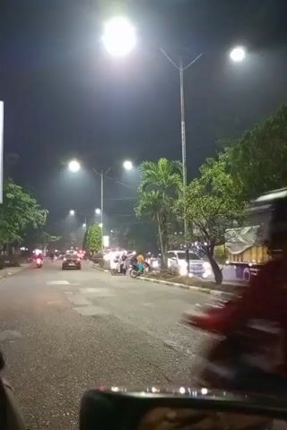 A short video clip shows a nighttime street scene, likely in a tropical or subtropical climate. The street is paved and appears wet, suggesting recent rain. Several streetlights illuminate the road, casting a bright glow and creating lens flares in the camera's view. Palm trees and other lush green foliage line the sides of the street. There is moderate traffic, primarily consisting of motorcycles and a few cars. A person on a motorcycle in the foreground is riding away from the camera. Other vehicles and pedestrians are visible further down the street. The overall atmosphere is one of a bustling, lively street at night.