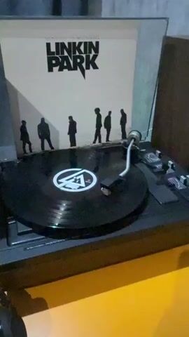 The video shows a close-up shot of a record player with a Linkin Park album, "Minutes to Midnight," displayed behind it. A black vinyl record with a white circular logo is spinning on the turntable. The record player is placed on a bright yellow surface, and the background is a textured gray wall. The lighting is warm and slightly diffused, highlighting the texture of the record sleeve and the details of the turntable. The camera is static, and there are no people or animals visible in the frame. The overall scene conveys a sense of vintage audio equipment and music appreciation.