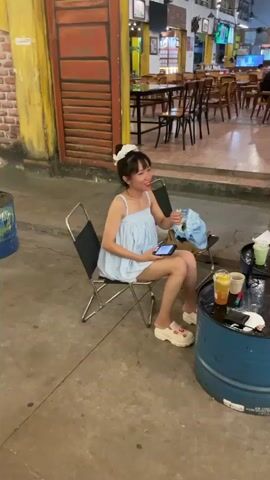 The video shows two separate scenes of a young woman dressed in different outfits standing outside what appears to be a restaurant. In the first scene, she is sitting on a silver metal chair wearing a light blue dress and white shoes, holding a smartphone in her hands. A blue bag rests on an adjacent chair, and a makeshift table made from a blue barrel holds drinks and other items. The background features the interior of the restaurant with tables and chairs. In the second scene, the woman is standing near a tree, wearing a dark blue dress and white shoes. She is smiling and appears to be posing. The environment features yellow and partially distressed walls, suggesting an older or rustic setting. The camera work is simple, likely captured handheld, focusing on the woman as the primary subject in both scenes.