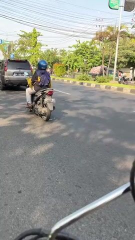 The video shows a person riding a white scooter on a paved road. The rider is wearing a blue jacket, dark pants, and a red helmet. They are accompanied by another person on the back of the scooter, who is wearing a dark top and patterned pants. In the background, there are trees, power lines, and a traffic light. Other vehicles, including a motorcycle and a car, are also visible on the road. The scene appears to be an urban or suburban environment during the daytime.