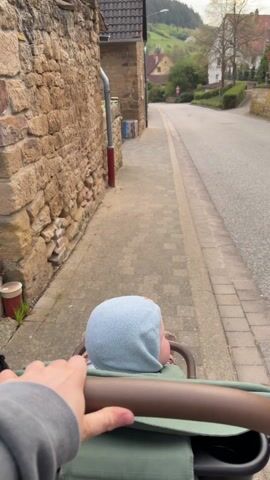 A person is pushing a baby in a stroller along a sidewalk next to a stone wall. The baby is wearing a light blue hat. The sidewalk runs parallel to a narrow road. The background shows houses and a hill with trees. The sky is overcast.