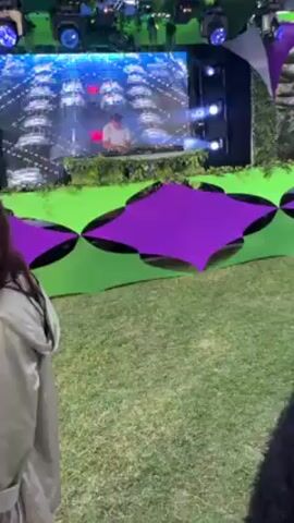 The video captures a lively scene at an outdoor music event. A DJ performs on a stage decorated with green and purple geometric patterns, backed by large video screens displaying abstract graphics. The partygoers, many holding drinks, are casually dressed.  Palm trees and resort buildings are visible in the background, suggesting a tropical location. The event features a canopy of green and purple fabric shapes overhead, enhancing the festive atmosphere.