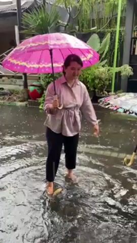 A woman and a child are standing in a flooded street. The woman holds a pink umbrella with a floral design. She is wearing a light-colored button-down shirt with a gathered waist, and black pants. She is barefoot. The child holds an umbrella with a black background and colorful floral design. She is wearing a light-colored dress, and appears to be barefoot. Both are smiling and appear to be enjoying themselves despite the water. The background includes houses, plants, and a black wrought-iron gate. It is raining.