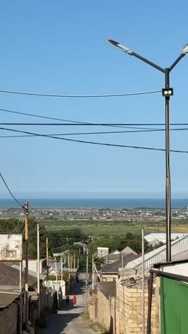 The video begins with a view of a street in a village, with houses and a street light visible. The street slopes downwards, and in the distance, there is a body of water, likely the sea, with a town or city along the coast. The sky is clear and blue. The camera then zooms out, offering a wider view of the landscape, including more houses, fields, and trees. The sea and the distant town remain visible. The video appears to be shot during the daytime, with bright sunlight. There are no distinct actions or interactions depicted. The overall environment is that of a coastal village or town with a scenic view of the sea.