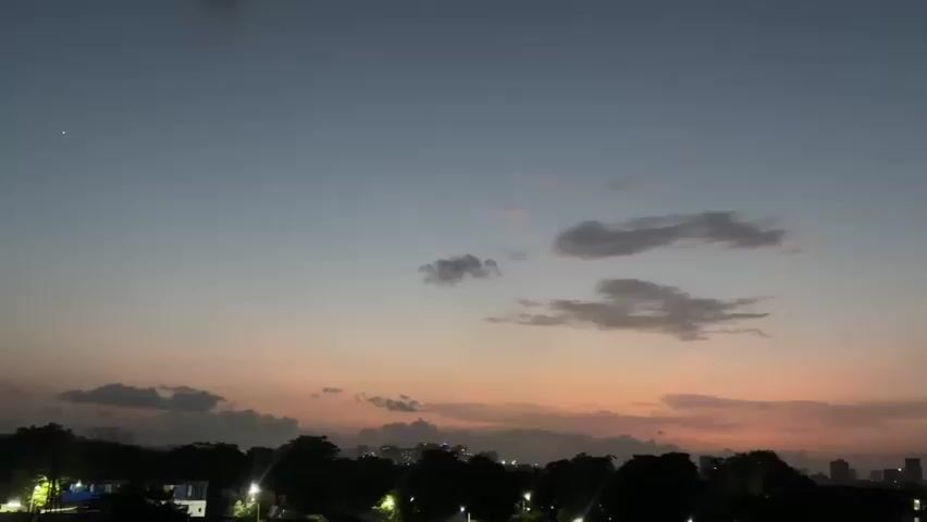 The video shows a time-lapse of a dusky sky with scattered clouds and a cityscape in the background. The sky transitions from a deep blue at the top to shades of orange and pink near the horizon, indicating either sunrise or sunset. A single bright star or planet is visible in the upper left portion of the sky. The cityscape below is silhouetted against the sky, with some lights visible, suggesting it is either early morning or late evening. The clouds are wispy and appear to be moving slowly across the sky. The overall impression is one of peaceful transition between day and night.