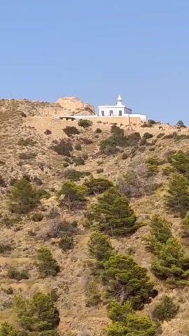A drone-like aerial view pans across a dry, scrub-covered hillside. On the crest of the hill, a white lighthouse stands against a clear blue sky. Below the hill, the ocean is visible, with a distant city skyline along the coast and a prominent rocky formation jutting out into the sea. The landscape is characterized by sparse, dry vegetation and scattered evergreen trees.