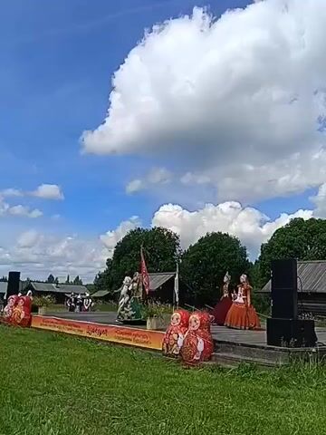 This is a wide shot of an outdoor stage performance on a sunny day with some clouds. On the stage, several people are dressed in traditional Russian folk costumes. Two people in green and white outfits are dancing in the center, while a person in a red and orange dress stands on the right side of the stage. In front of the stage, two large matryoshka dolls are displayed. The background consists of green grass, trees, and some wooden buildings, suggesting a rural or historical setting. Two large black speakers are placed on the right side of the stage. The sky is blue with fluffy white clouds. The overall atmosphere appears festive.