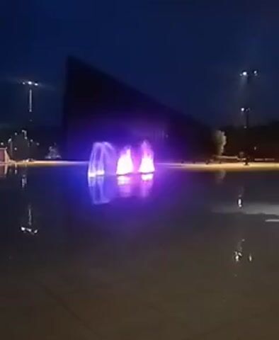 The video shows an outdoor fountain at night. The fountain water is illuminated with purple lights and reflects in the large, shallow pool of water. In the background, a dark, angular building is visible, and streetlights can be seen in the distance. The sky is dark, indicating it is nighttime. There are no people clearly visible in the frame.