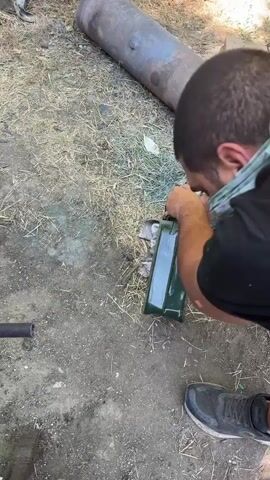 A person is seen from behind, wearing a black t-shirt and a green bandana around their neck, kneeling outdoors on a dirt and dry grass surface. The person is holding a green gas can and appears to be wiping off some blue substance from it with a grey cloth. In the background, there is a large, rusty cylindrical object lying on the ground, possibly a tank or pipe. Scattered around are other items including a red and silver can, some tools, and a pair of black sneakers. The scene suggests some sort of outdoor work or repair activity.